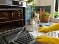Hände mit gelben Handschuhen reinigen den Ofenrost mit einem blauen Schwamm.