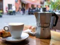 Ein Espresso-Kocher steht neben einer Tasse Cappuccino und einem Croissant auf einem Holztisch. Nach dem Genuss ist das espressokocher reinigen angesagt.