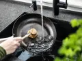 Eine Hand schrubbt mit einer Bürste eine Gusseisenpfanne unter fließendem Wasser. So gelingt das Gusseisenpfanne reinigen mühelos.
