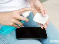 Hände desinfizieren: Eine Person sprüht Desinfektionsmittel auf ein Wattepad, um das Handy zu reinigen.
