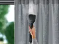 Hand hält eine AEG Dampfbürste, um graue Vorhänge zu glätten. Dampf steigt auf.