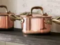 Drei glänzende Kupfertopf-Sets mit Messinggriffen stehen auf einem dunklen Regal. Sie warten darauf, mit einem speziellen Mittel zum Kupfer reinigen wieder zum Strahlen gebracht zu werden.