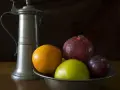 Ein Zinnkrug steht neben einer Schale mit Früchten: Orange, Apfel, Granatapfel und Pflaume. Zeit für zinn reinigen.