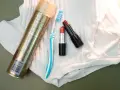 Haarspray, Zahnbürste und Lippenstifte liegen auf weißem Stoff. Ein Tipp, wie man Lippenstift aus Kleidung entfernen kann.