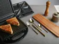 Zwei goldbraune Sandwiches liegen im geöffneten Sandwichmaker. Daneben liegen Besteck, ein Holzbrett und Gewürzmühlen. Zeit, den Sandwichmaker zu reinigen.