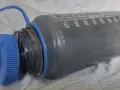 Graue Trinkflasche mit blauer Kappe, die eine gründliche Reinigung benötigt. Messskala bis 1000 ml sichtbar.