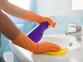 Hände in orangen Handschuhen sprühen Reinigungsmittel auf ein Waschbecken und wischen mit einem gelben Putzlappen.
