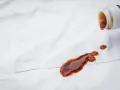 Braune Flüssigkeit tropft aus einer Flasche auf weiße Kleidung. Tipps zur **balsamico flecken entfernen** sind hier gefragt.