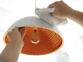 Hände reinigen einen modernen Lampenschirm mit einem weißen Tuch. Das Innere des Schirms ist orange und geriffelt.