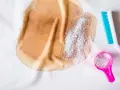Brauner Fleck auf weißem Stoff, dane Waschpulver in einem rosa Löffel und eine blaue Bürste. So lässt sich **Haarfarbe aus Kleidung entfernen**.