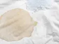 Ein brauner Fleck auf weißem Stoff, dane ein Haufen Waschpulver. So lassen sich hartnäckige teeflecken entfernen.