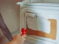 Eine Hand mit einem Farbroller trägt weiße Farbe auf ein Holzmöbel auf. Das Holz wird vorbereitet, um es zu entfetten und neu zu streichen.