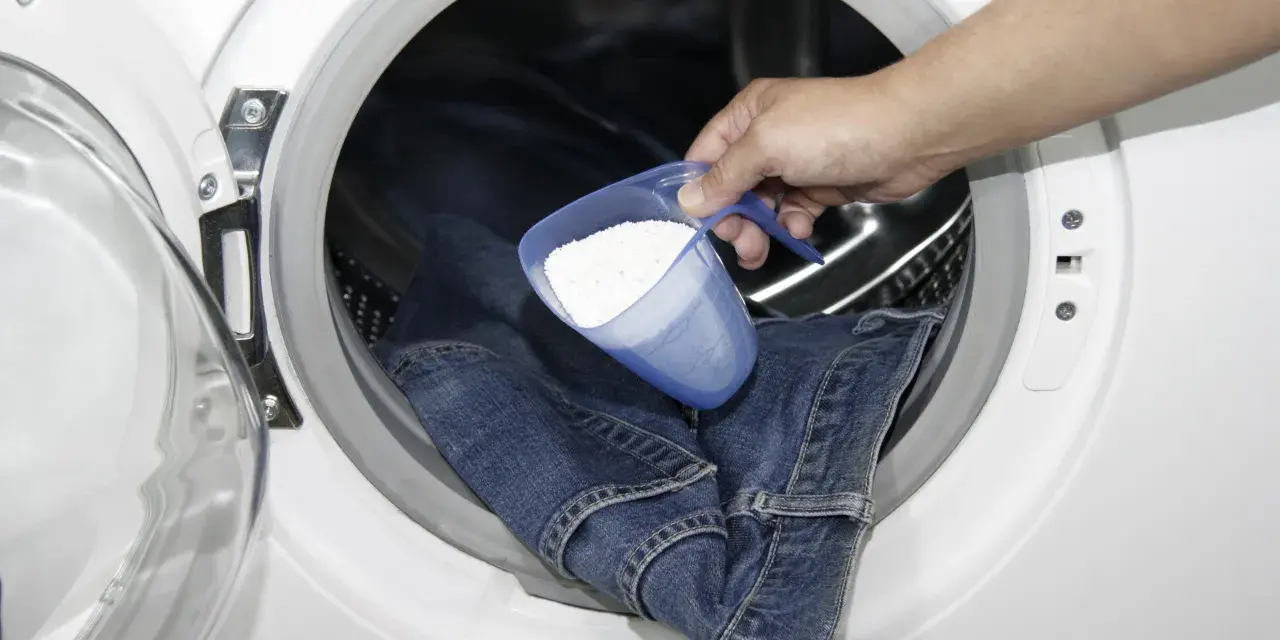 Eine Hand gibt Waschpulver in eine Waschmaschine, um Kettenfett aus Jeans zu entfernen.