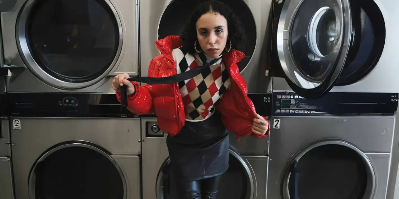 Junge Frau in roter Daunenjacke und Lederrock vor Waschmaschinen. Sie hält ein schwarzes Band. Kunstleder waschen erfordert Vorsicht.