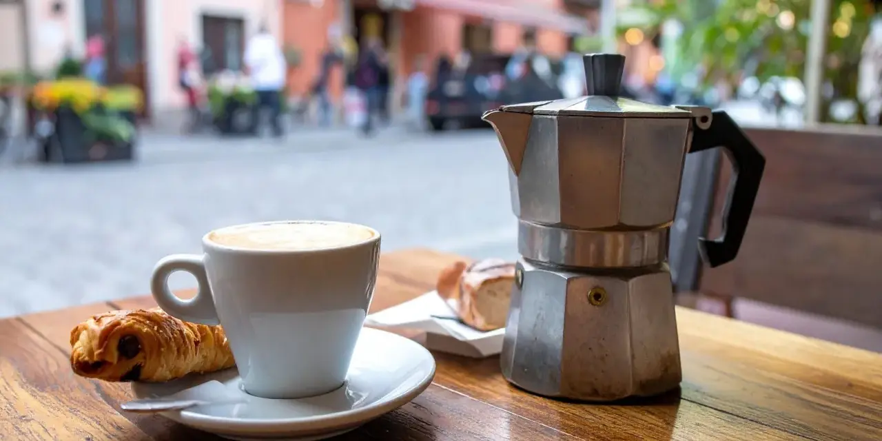 Ein Espresso-Kocher steht neben einer Tasse Cappuccino und einem Croissant auf einem Holztisch. Nach dem Genuss ist das espressokocher reinigen angesagt.