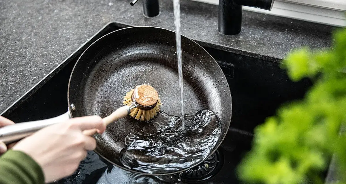 Eine Hand schrubbt mit einer Bürste eine Gusseisenpfanne unter fließendem Wasser. So gelingt das Gusseisenpfanne reinigen mühelos.