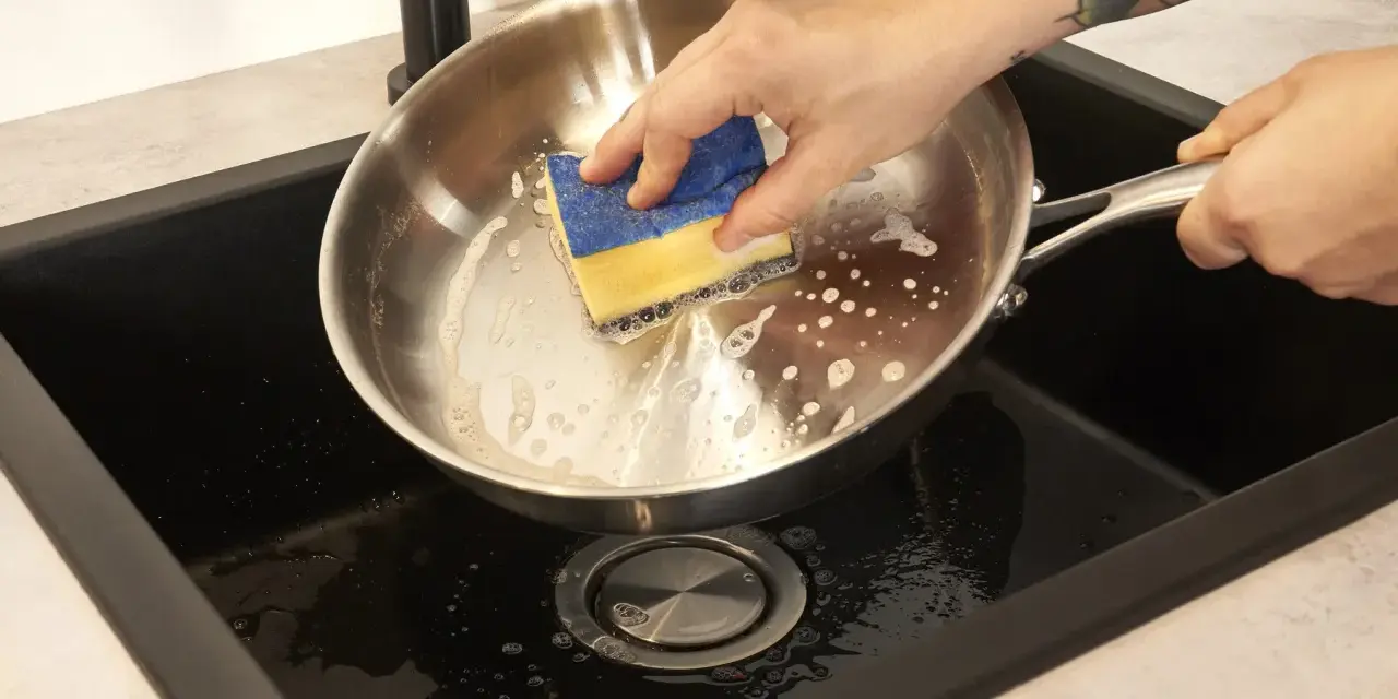 Hand reinigt mit einem Schwamm den schmutzigen Pfannenboden in der Spüle.