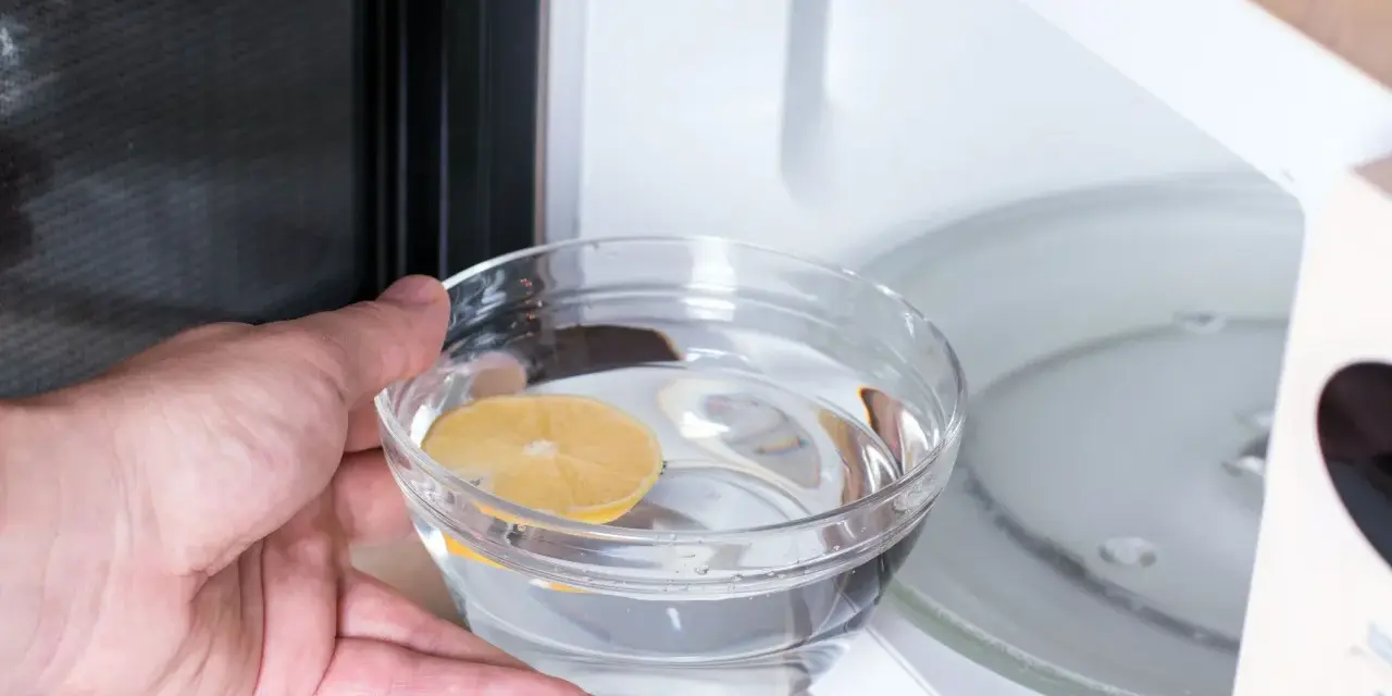 Hand placing a bowl of water with lemon slice into a microwave for cleaning.