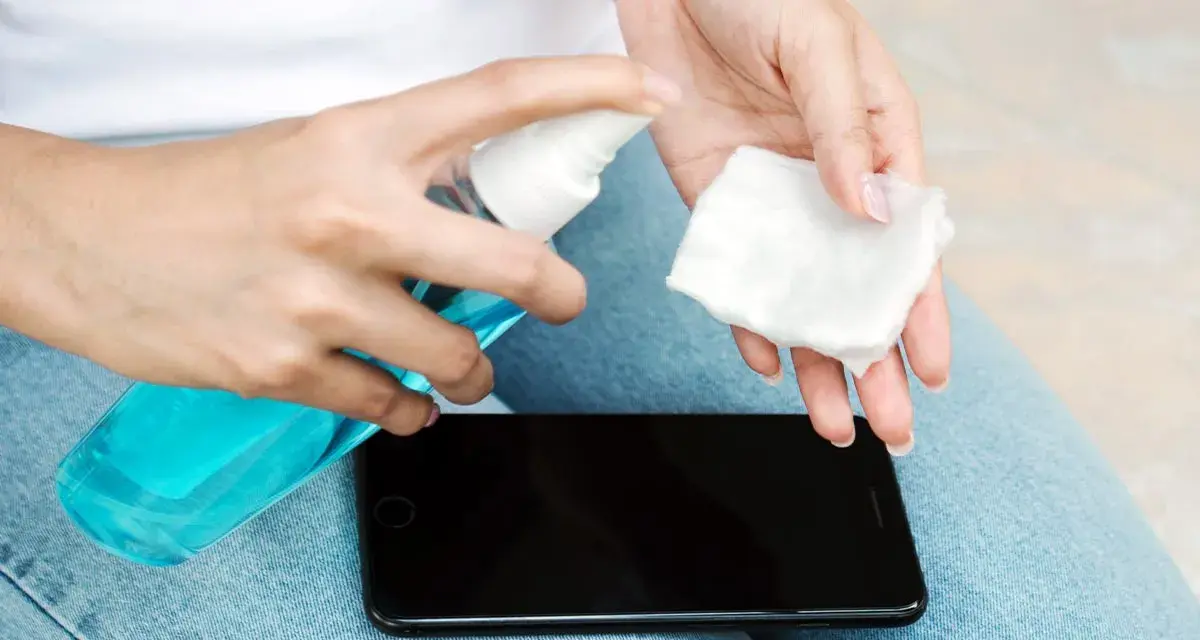 Hände desinfizieren: Eine Person sprüht Desinfektionsmittel auf ein Wattepad, um das Handy zu reinigen.