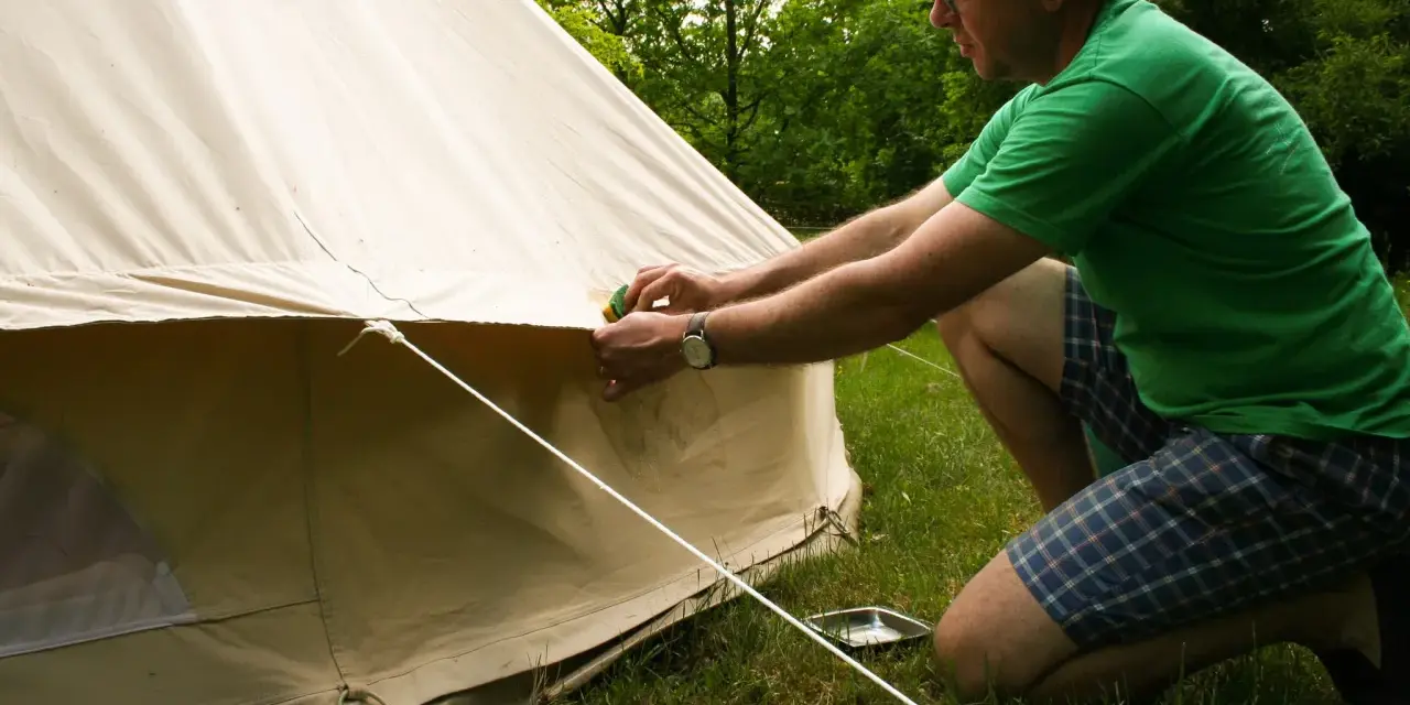 Mann reinigt mit einem Schwamm das beige Zelt. Er kniet auf dem Gras neben dem Zelt.