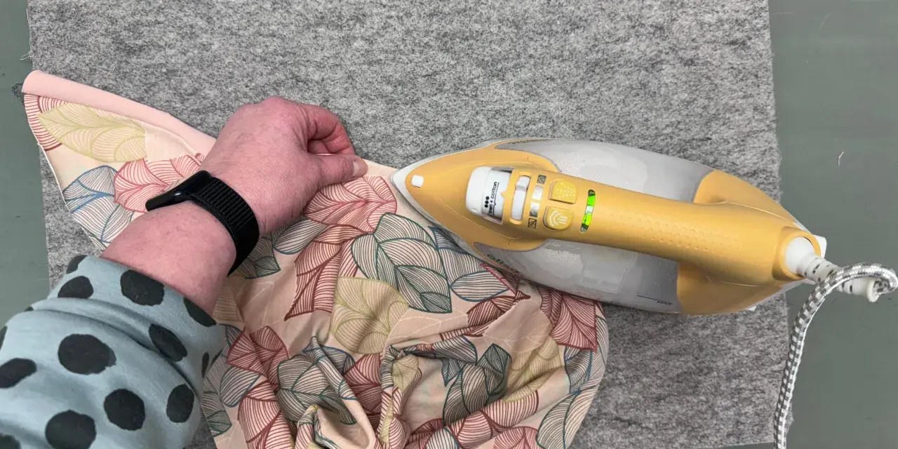 Ein kleines Bügeleisen zum Nähen glättet Stoff mit Blattmuster auf einer Filzmatte.