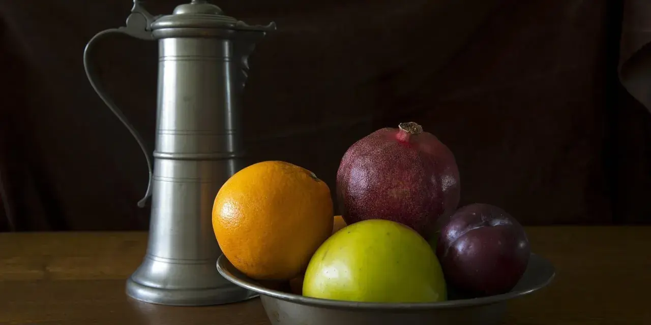Ein Zinnkrug steht neben einer Schale mit Früchten: Orange, Apfel, Granatapfel und Pflaume. Zeit für zinn reinigen.