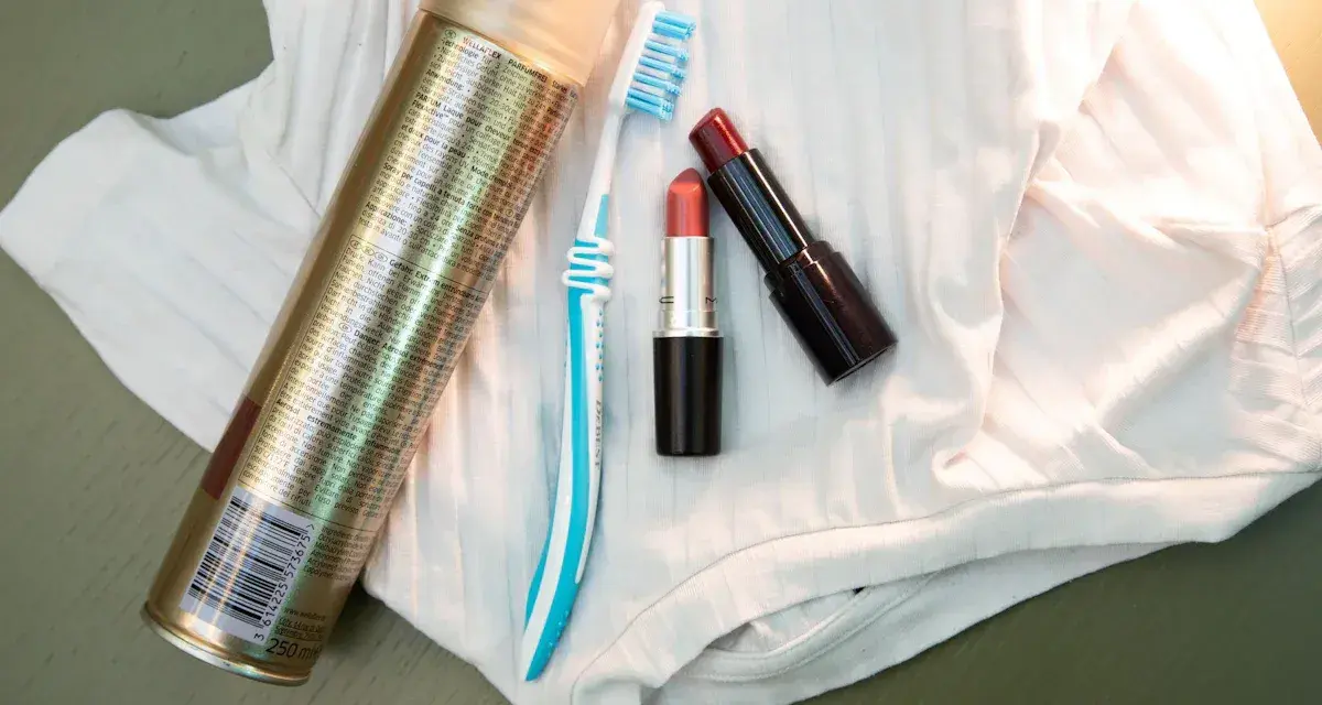 Haarspray, Zahnbürste und Lippenstifte liegen auf weißem Stoff. Ein Tipp, wie man Lippenstift aus Kleidung entfernen kann.