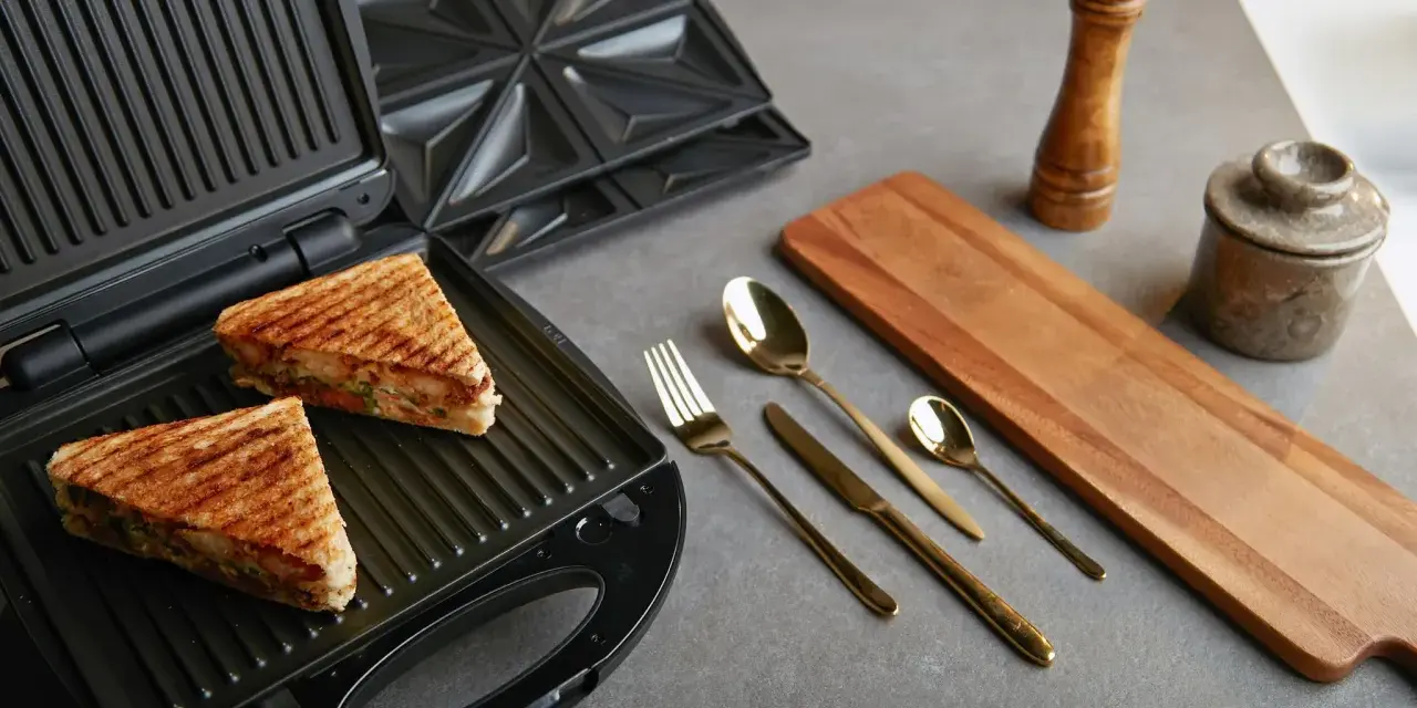 Zwei goldbraune Sandwiches liegen im geöffneten Sandwichmaker. Daneben liegen Besteck, ein Holzbrett und Gewürzmühlen. Zeit, den Sandwichmaker zu reinigen.