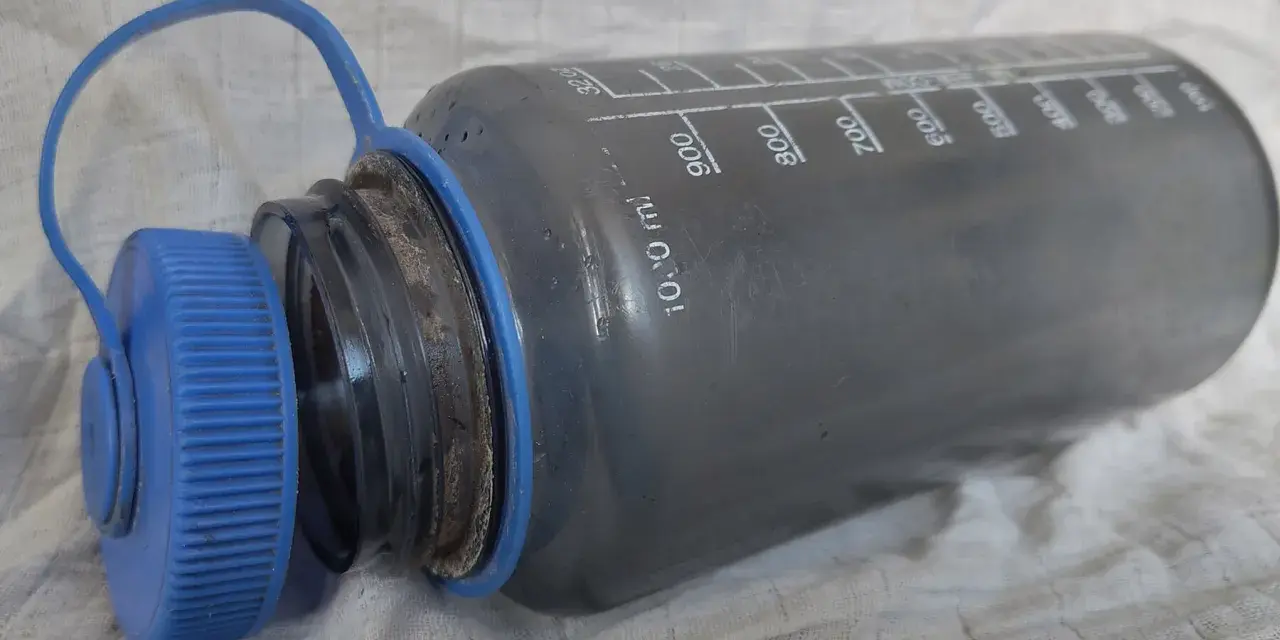 Graue Trinkflasche mit blauer Kappe, die eine gründliche Reinigung benötigt. Messskala bis 1000 ml sichtbar.
