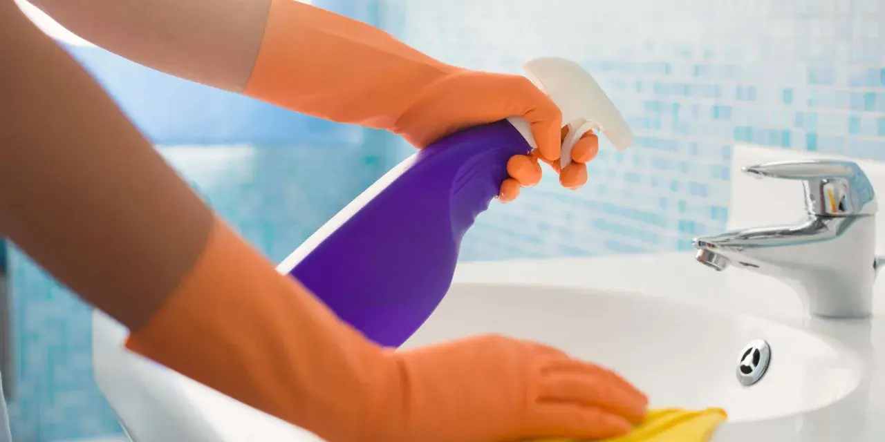 Hände in orangen Handschuhen sprühen Reinigungsmittel auf ein Waschbecken und wischen mit einem gelben Putzlappen.