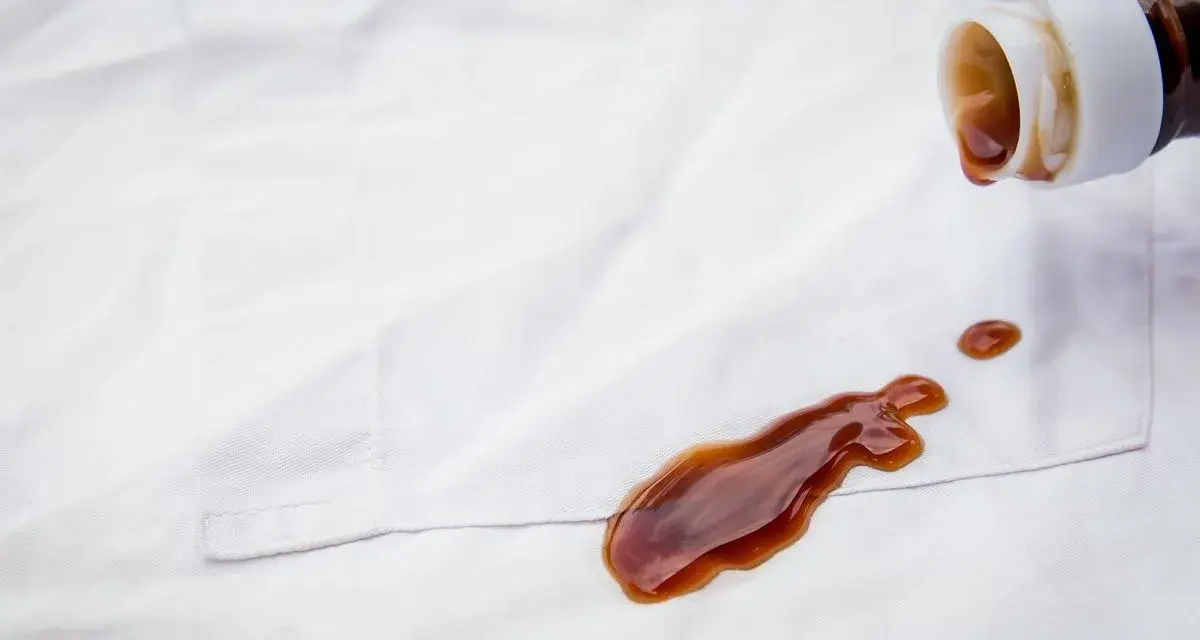 Braune Flüssigkeit tropft aus einer Flasche auf weiße Kleidung. Tipps zur **balsamico flecken entfernen** sind hier gefragt.