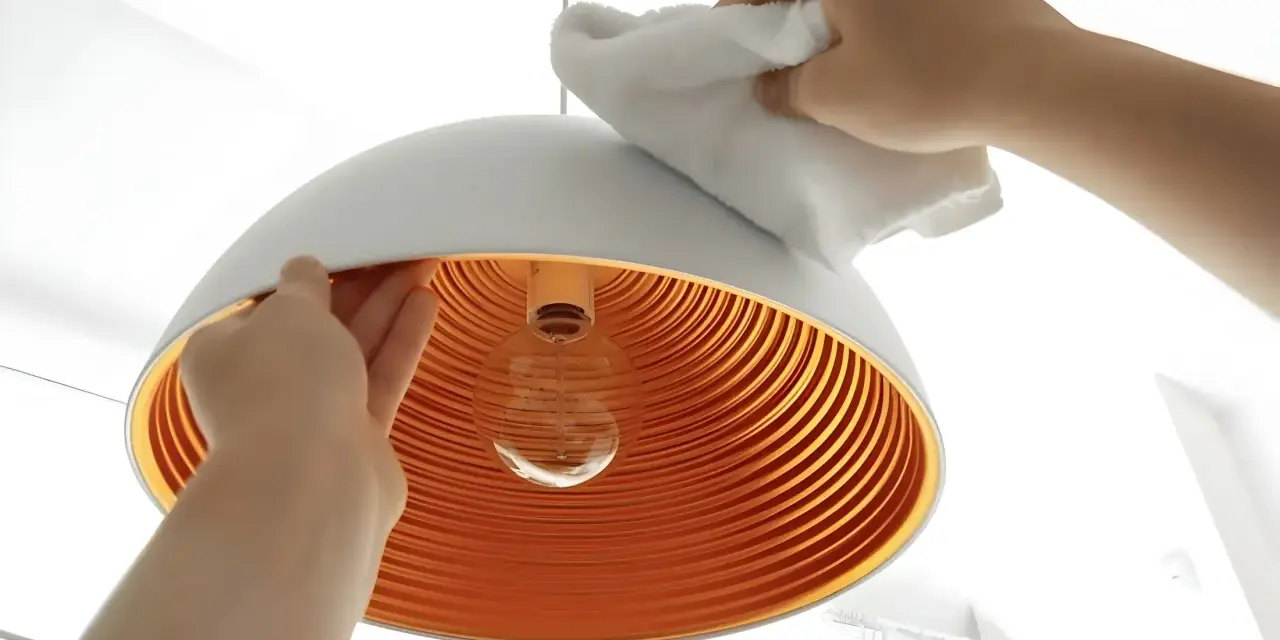 Hände reinigen einen modernen Lampenschirm mit einem weißen Tuch. Das Innere des Schirms ist orange und geriffelt.