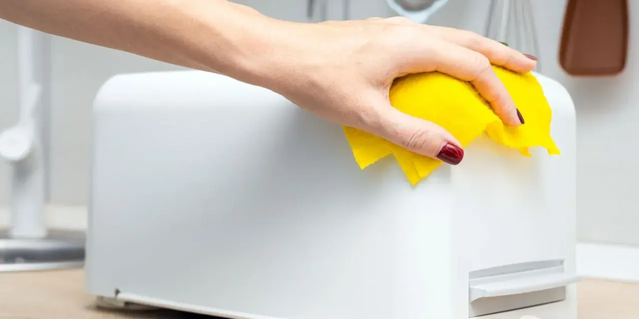 Eine Hand mit gelbem Tuch wischt über einen weißen Toaster. So einfach kann toaster reinigen sein.