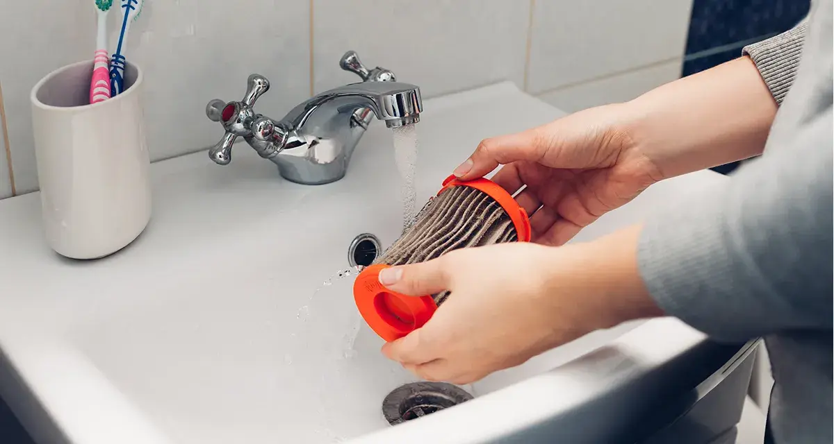 Hände reinigen einen staubigen Staubsaugerfilter unter fließendem Wasser im Waschbecken.