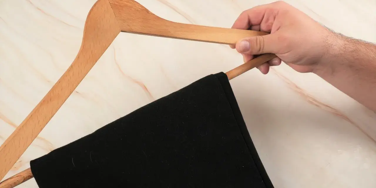 Hand hält einen Holz-Kleiderbügel mit einer schwarzen Anzughose. Zeit, die Anzughose zu waschen.