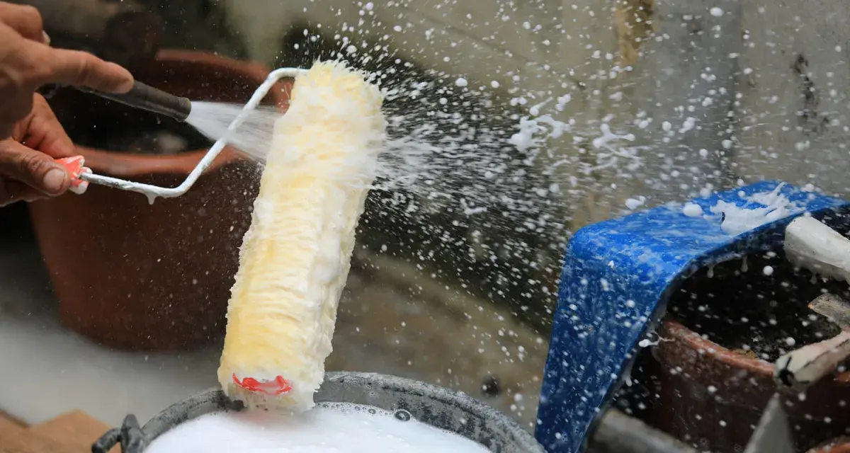 Eine Hand spritzt Wasser auf eine Malerrolle, um sie in einem Eimer mit Seifenwasser zu reinigen.
