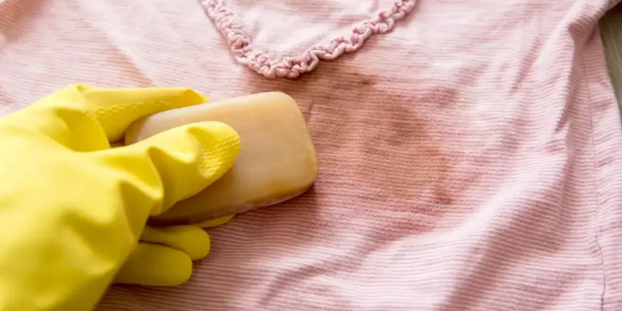 Gelbe Handschuhe halten Gallseife, um einen Fleck auf einem rosafarbenen gestreiften Shirt zu behandeln.