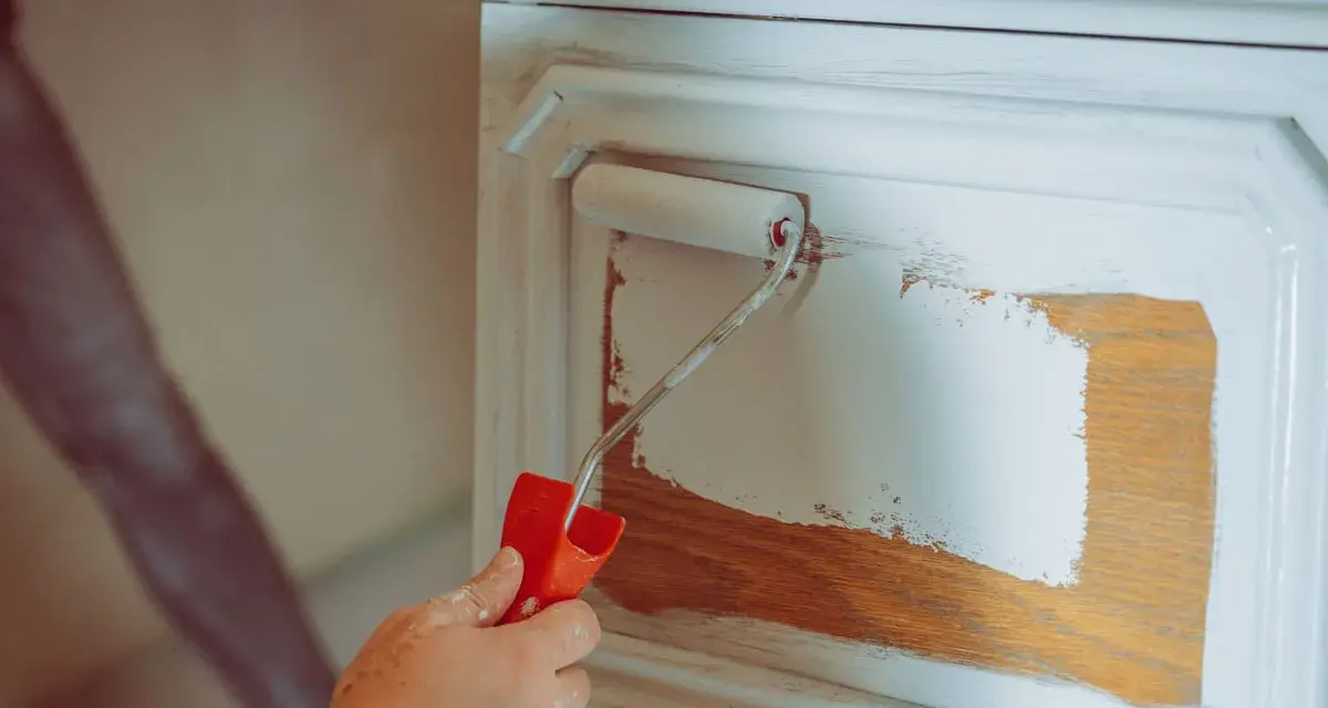 Eine Hand mit einem Farbroller trägt weiße Farbe auf ein Holzmöbel auf. Das Holz wird vorbereitet, um es zu entfetten und neu zu streichen.