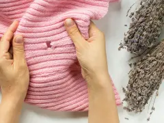 Hände prüfen ein rosafarbenes Strickteil auf Löcher, ein Zeichen, dass man Kleidermotten erkennen muss. Lavendelzweige liegen daneben.