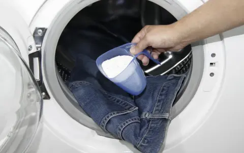 Eine Hand gibt Waschpulver in eine Waschmaschine, um Kettenfett aus Jeans zu entfernen.