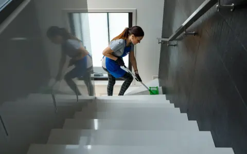 Frau wischt Treppenstufen mit einem Mopp. Sie trägt eine Schürze und Handschuhe. Man fragt sich, wie oft wischen nötig ist, um diesen Glanz zu erhalten.