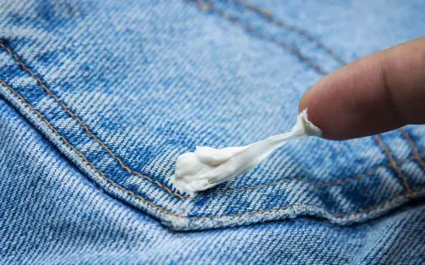 Finger versucht, Kaugummi aus Jeans zu entfernen. Der klebrige weiße Klumpen haftet an der blauen Denim-Oberfläche.