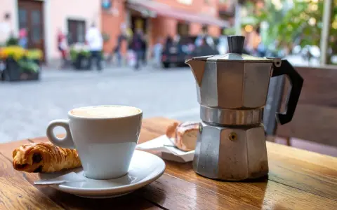 Ein Espresso-Kocher steht neben einer Tasse Cappuccino und einem Croissant auf einem Holztisch. Nach dem Genuss ist das espressokocher reinigen angesagt.