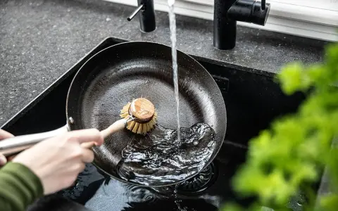Eine Hand schrubbt mit einer Bürste eine Gusseisenpfanne unter fließendem Wasser. So gelingt das Gusseisenpfanne reinigen mühelos.