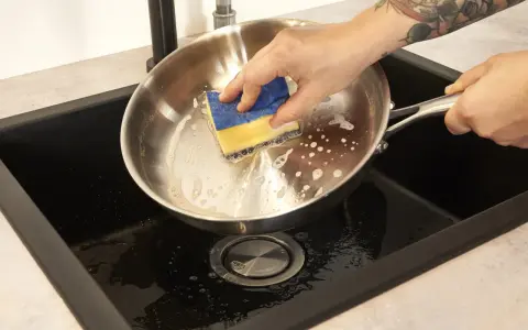 Hand reinigt mit einem Schwamm den schmutzigen Pfannenboden in der Spüle.