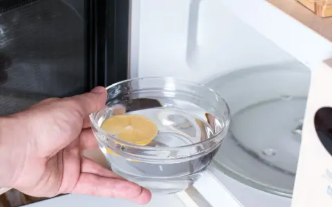 Hand placing a bowl of water with lemon slice into a microwave for cleaning.