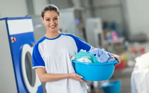 Lächelnde Frau mit Wäschekorb voller bunter Kleidung vor einer Waschmaschine. Sie weiß, wie man richtig waschen.