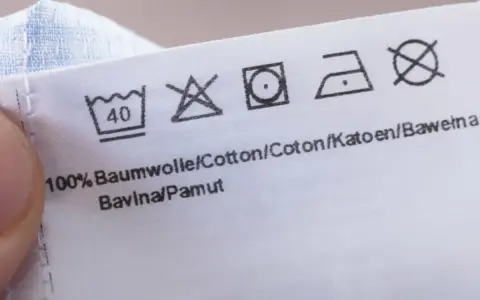 Pflegesymbol: Nicht im Trockner trocknen. Das Etikett zeigt auch Wasch- und Bügelsymbole für 100% Baumwolle.