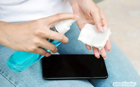 Hände desinfizieren: Eine Person sprüht Desinfektionsmittel auf ein Wattepad, um das Handy zu reinigen.