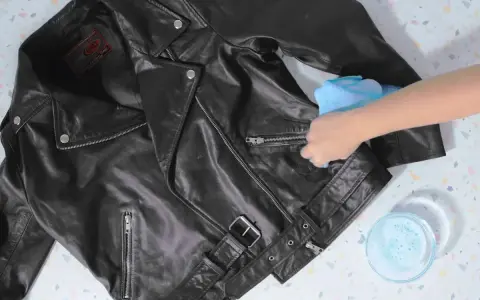 Schwarze Lederjacke reinigen mit einem feuchten Tuch. Eine Hand wischt über die Jacke, daneben eine Schale mit Seifenwasser.
